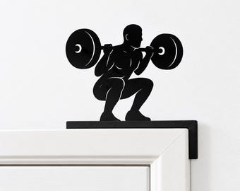 Könnte beinhalten: Ein Türaufsatz aus schwarzem Metall mit der Silhouette einer Person, die eine Kniebeuge mit einer Langhantel ausführt. Die Figur befindet sich in einer tiefen Hocke und hält eine Hantel mit großen runden Gewichtsscheiben auf den Schultern, montiert auf einem L-förmigen Halter.