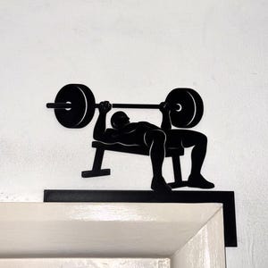 Peut inclure: Silhouette noire en métal d'une personne faisant du développé couché avec une barre. La silhouette est allongée sur un banc, la barre au-dessus de sa poitrine. L'œuvre est fixée sur un mur blanc et un cadre noir.