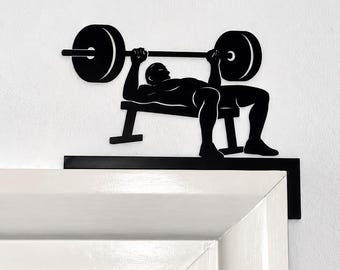 Könnte beinhalten: Ein Türschild aus schwarzem Metall mit der Silhouette einer Person, die eine Bankdrückübung mit einer Langhantel ausführt. Das Dekorationselement ist so gestaltet, dass es auf der oberen Ecke eines Türrahmens platziert werden kann.
