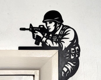Puede incluir: Silueta metálica negra de un soldado apuntando con un rifle. La obra de arte está montada en la esquina de un marco de puerta blanco. El soldado lleva casco y equipo militar.