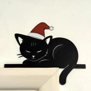 Puede incluir: Decoración con forma de gato negro con un gorro de Papá Noel rojo y blanco. El gato está tumbado con los ojos cerrados. La decoración está colocada sobre una superficie blanca, ideal para la decoración navideña.
