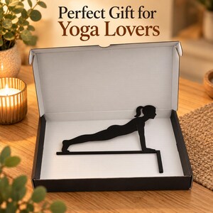 K&ouml;nnte beinhalten: Eine schwarze Metallsilhouette einer Person in Yoga-Plankenposition, montiert auf einer horizontalen Stange mit vertikaler St&uuml;tze, pr&auml;sentiert in einer offenen wei&szlig;-schwarzen Pappschachtel. Der Text Perfect Gift for Yoga Lovers ist oberhalb der Box zu sehen.