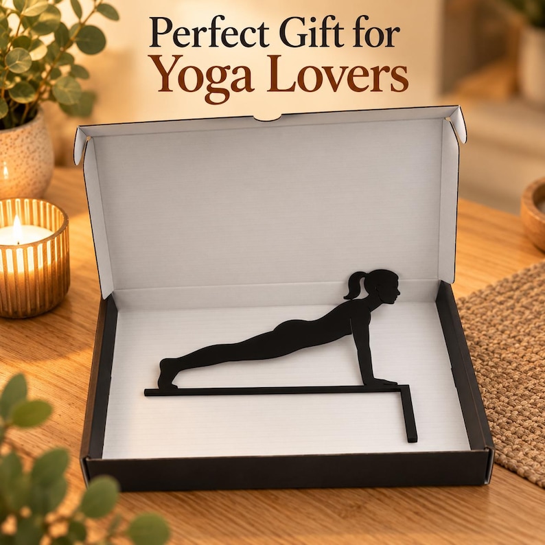 K&ouml;nnte beinhalten: Eine schwarze Metallsilhouette einer Person in Yoga-Plankenposition, montiert auf einer horizontalen Stange mit vertikaler St&uuml;tze, pr&auml;sentiert in einer offenen wei&szlig;-schwarzen Pappschachtel. Der Text Perfect Gift for Yoga Lovers ist oberhalb der Box zu sehen.