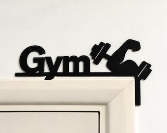 Puede incluir: Un letrero de esquina para puerta en metal negro que presenta la palabra Gym en una fuente sans-serif negrita, acompañada por la silueta de un brazo flexionado sosteniendo una mancuerna, diseñado para el marco de la puerta.