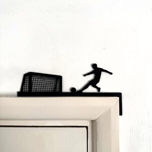 May include: Black metal wall decoration depicting a football player kicking a ball towards a goal. The silhouette design is mounted on a white surface, creating a contrast. The goal has a net.