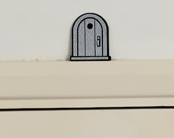 Puede incluir: Una pequeña pegatina de puerta de madera arqueada con un contorno negro. La puerta está pintada de gris claro con detalles de vetas de madera, un pomo negro redondo y un tirador rectangular. La pegatina está colocada sobre una superficie blanca.
