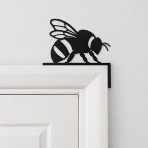 Può includere: Una sagoma di ape in metallo nero progettata per essere posizionata sull'angolo superiore di una cornice di porta. L'ape presenta dettagli intagliati sulle ali e strisce sul corpo, montata su un supporto a L che poggia sulla modanatura.