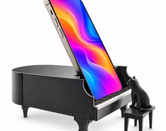 Puede incluir: Un soporte para smartphone con forma de piano de cola en miniatura negro, que incluye una pequeña figura de gato negro sentada en un banco de piano a juego. El soporte sostiene un smartphone con una pantalla de colores vivos.