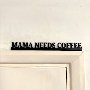 Könnte beinhalten: Ein schwarzes Metallschild mit der Aufschrift "MAMA NEEDS COFFEE" über einer weißen Tür. Das Schild ist in fetter Schrift gehalten und an einer weißen Wand befestigt. Der Türrahmen ist cremefarben gestrichen.
