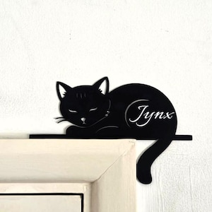 Puede incluir: Decoración de pared con forma de gato negro con el nombre "Jynx" en fuente cursiva. El gato está acostado, con los ojos cerrados. La decoración está montada en una repisa negra sobre un marco de puerta blanco.