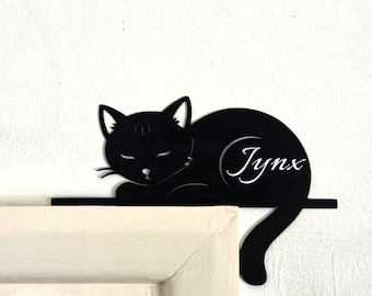 Puede incluir: Decoración de pared con forma de gato negro con el nombre "Jynx" en fuente cursiva. El gato está acostado, con los ojos cerrados. La decoración está montada en una repisa negra sobre un marco de puerta blanco.