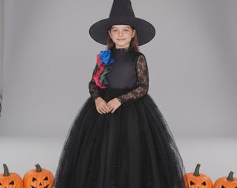 May include: A young person in a black witch costume with a tall, pointed hat and a long, flowing skirt. The dress features lace sleeves and a colorful floral accent. Orange jack-o'-lanterns are in the foreground, creating a Halloween theme.