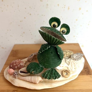 May include: A green frog made from seashells with googly eyes sitting on a bed of seashells on a wooden base.