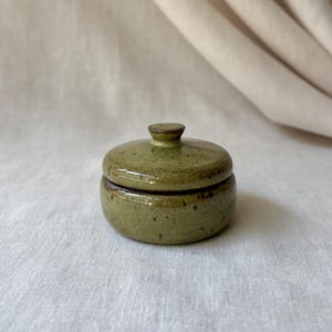May include: A small, round ceramic lidded jar featuring a speckled olive green glaze. The container has a smooth, cylindrical body and a matching lid with a small, centered knob handle. The surface displays dark brown speckles throughout the finish.