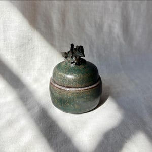 May include: A small, round, dark green ceramic jar with a matching lid. The surface features a mottled, speckled texture with brown undertones. The lid is topped with a jagged, dark, sculptural handle that resembles a rough, organic form.