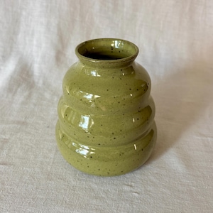 May include: A small, olive green ceramic vase with a glossy finish and dark speckles. The vase features a distinct three-tiered, rounded beehive-like shape with a narrow neck and a wide, circular opening at the top, showcasing a smooth, glazed texture.