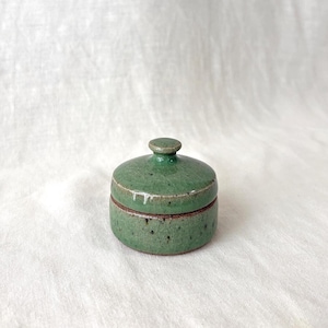 May include: A small, round, lidded ceramic jar with a glossy green glaze featuring dark speckles throughout. The lid has a central knob handle, and the base of the jar shows a thin, exposed brown rim where the glaze ends, creating a distinct contrast.