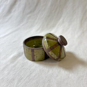 May include: A small, open ceramic jar with a lid. The jar and lid are olive green with brown geometric designs. The lid has a small, round, brown knob. The interior of the jar is a darker green.
