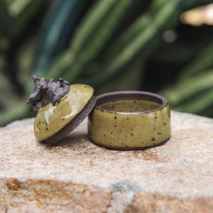 May include: A small, round ceramic container with a glossy olive-green glaze and dark brown speckles. The lid features a textured, dark brown, organic-shaped handle that resembles a piece of driftwood or a gnarled twig, contrasting with the smooth base.