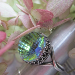 May include: A silver ring with a large, iridescent green and blue gemstone. The gemstone has a faceted, almost teardrop shape.