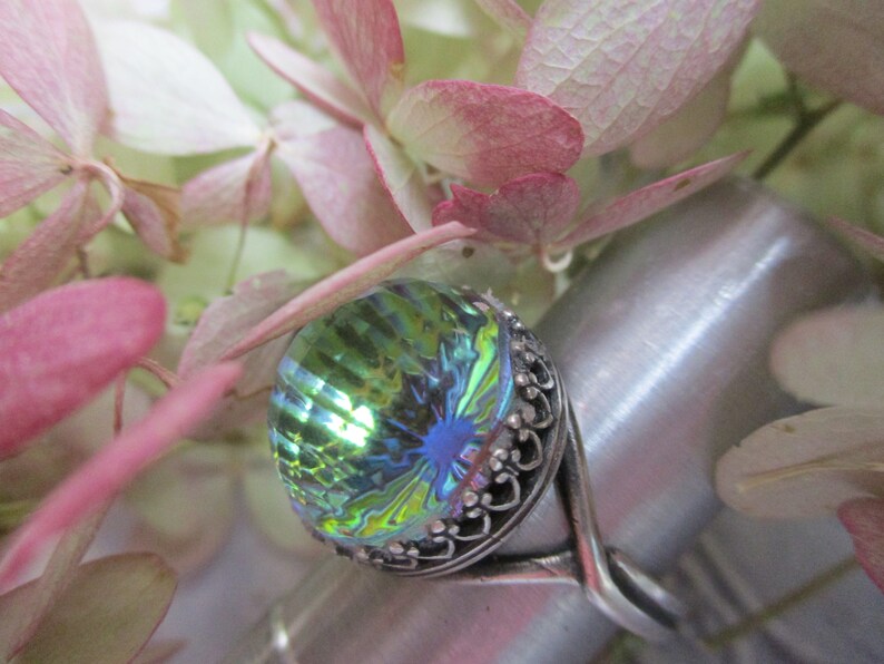 May include: A silver ring with a large, iridescent green and blue gemstone. The gemstone has a faceted, almost teardrop shape.