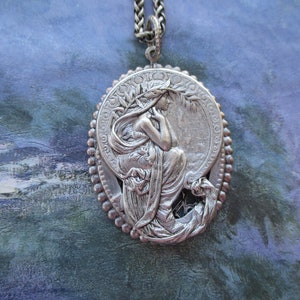 May include: A silver pendant necklace featuring a detailed relief of a woman with flowing hair and drapery, set within a circular frame with a beaded border.