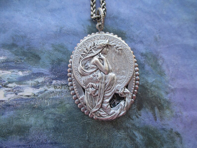May include: A silver pendant necklace featuring a detailed relief of a woman with flowing hair and drapery, set within a circular frame with a beaded border.