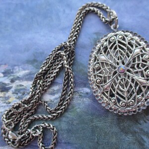 May include: A silver chain necklace with a large oval pendant. The pendant is made of intricate filigree work and features a small iridescent stone in the center.