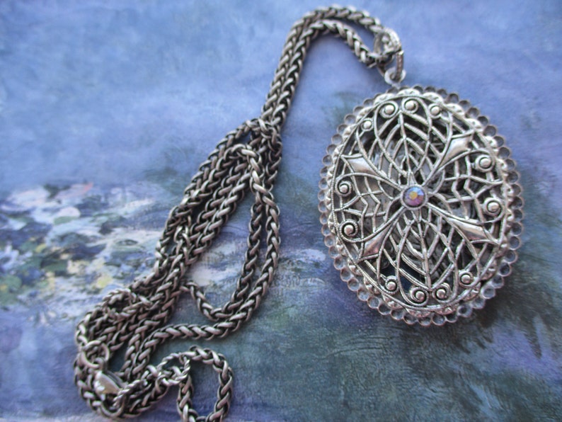 May include: A silver chain necklace with a large oval pendant. The pendant is made of intricate filigree work and features a small iridescent stone in the center.
