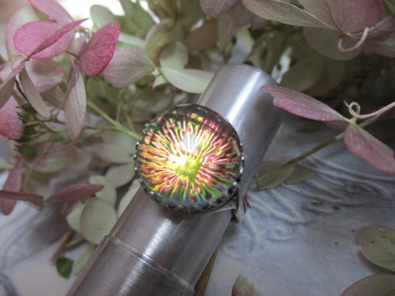 May include: A silver ring with a round iridescent glass stone. The stone has a multi-colored, swirling pattern that resembles a flower.