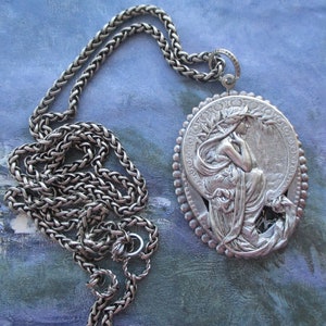 May include: A silver chain necklace with a large, oval pendant featuring a detailed, sculpted image of a woman with flowing hair and drapery. The pendant is surrounded by a decorative border of small beads.