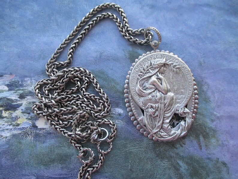 May include: A silver chain necklace with a large, oval pendant featuring a detailed, sculpted image of a woman with flowing hair and drapery. The pendant is surrounded by a decorative border of small beads.