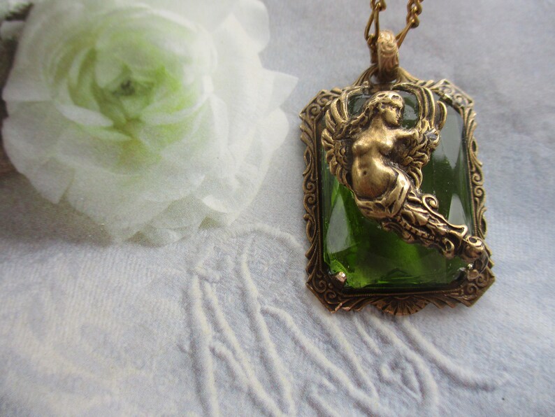 K&ouml;nnte beinhalten: Eine goldene Halskette mit einem gr&uuml;nen Glas-Cameo, das eine Meerjungfrau mit Fischschwanz und Fl&uuml;geln zeigt. Der Cameo ist in einen goldenen Rahmen mit verzierten Details gefasst.