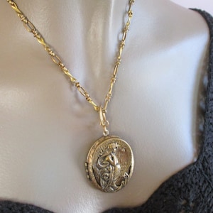 May include: A gold chain necklace with a gold locket pendant featuring a detailed design of a woman with flowing hair and a floral motif.