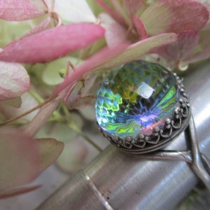May include: A silver ring with a large iridescent green, blue, and yellow glass stone set in a decorative silver bezel. The ring is on a silver display stand.