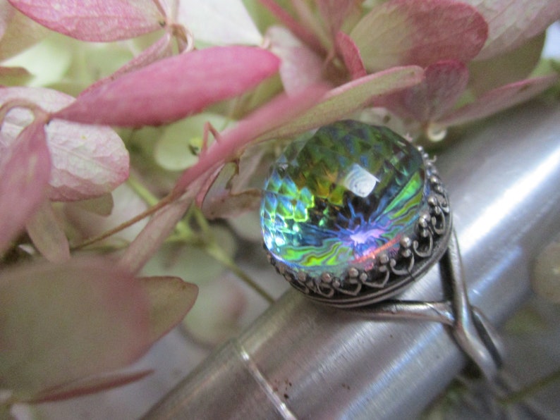 May include: A silver ring with a large iridescent green, blue, and yellow glass stone set in a decorative silver bezel. The ring is on a silver display stand.