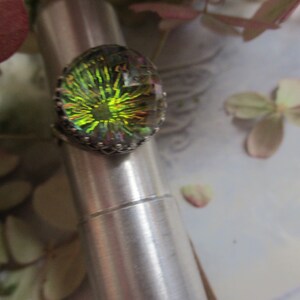 May include: A silver ring with a large oval-shaped iridescent gemstone. The gemstone has a green, yellow, and blue color with a starburst pattern.