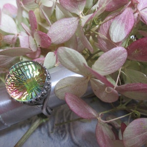 May include: A silver ring with a round iridescent green and pink glass stone set in a silver bezel. The ring is on a silver display stand.