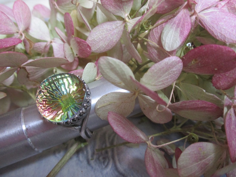 May include: A silver ring with a round iridescent green and pink glass stone set in a silver bezel. The ring is on a silver display stand.