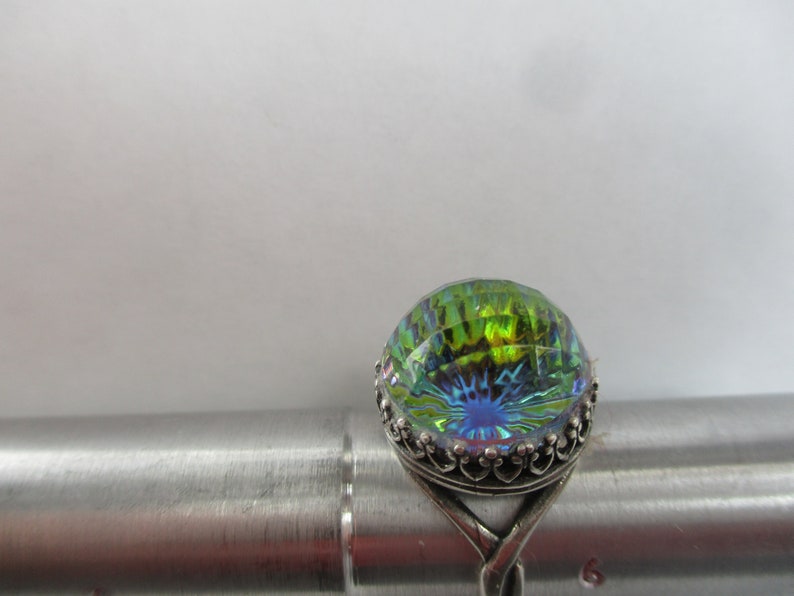 May include: A silver ring with a large iridescent green and blue gemstone. The gemstone is faceted and has a starburst pattern.