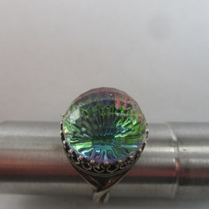 May include: A silver ring with a large, round, iridescent green and blue gemstone. The gemstone has a faceted design and is set in a silver band with a decorative border.