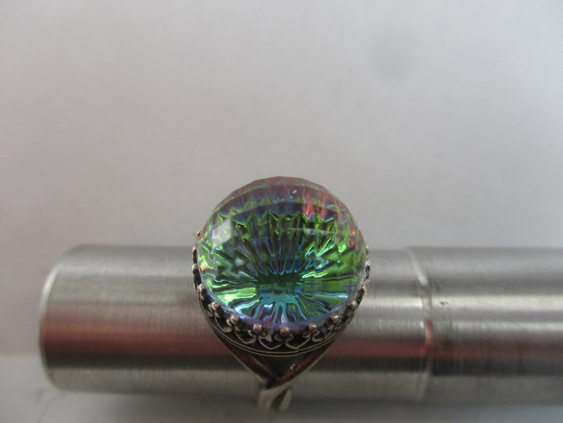 May include: A silver ring with a large, round, iridescent green and blue gemstone. The gemstone has a faceted design and is set in a silver band with a decorative border.