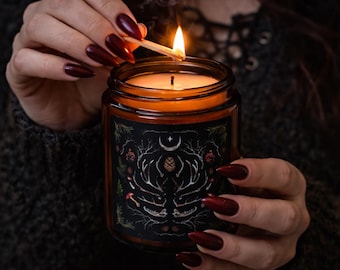 May include: A lit candle in a glass jar with a black label featuring a white antler design, a crescent moon, and botanical elements. The candle is being lit with a match. The jar is held by a person with dark red nail polish.