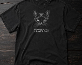 May include: Black t-shirt featuring a detailed illustration of a cat's face with piercing white eyes. The text "BEHIND THIS FACE LIES VIOLENCE" is printed below the cat's face. The shirt is made of a soft material.