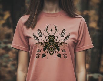 May include: A dusty rose t-shirt featuring a detailed illustration of a beetle with antlers and wings, surrounded by botanical elements. The design is in shades of black and gold. The shirt is a short-sleeved crew neck.