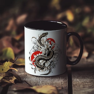 May include: A white ceramic mug with a black rim and handle. The mug features a black and white illustration of a snake wrapped around a flower with red and white geometric shapes in the background.