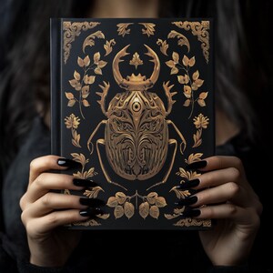 May include: A black hardcover journal with a gold beetle design. The beetle is intricately detailed with ornate patterns and a single eye in the center. The background is black with gold floral accents.