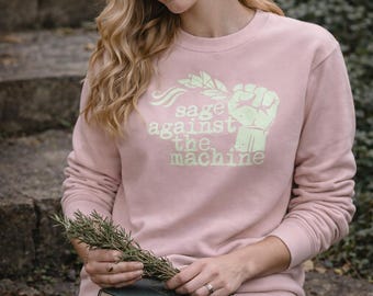 May include: A light pink sweatshirt with the text "sage against the machine" in a light green color. The sweatshirt has long sleeves and a crew neck. The image also shows a book and a sprig of rosemary.
