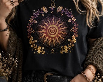 May include: Black t-shirt featuring a sun design with a spiral center and radiating lines. The sun is surrounded by a floral wreath with a bee and star accents. The design incorporates shades of gold, purple, and orange.