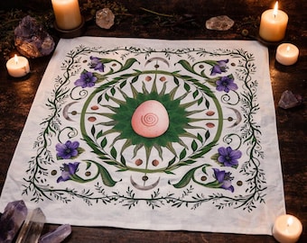May include: A white cloth with a circular design featuring a pink egg-shaped stone at the center, surrounded by a green sunburst, purple flowers, and crescent moons. Lit candles and crystals surround the cloth, creating a mystical ambiance.